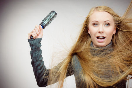 Blonde woman styling her very long blonde hair. Astonished girl with blowing messy hair holding round hairbrush. Haircare.の写真素材