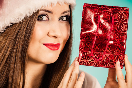 Smiling woman is happy to give Christmas gifts. Female wearing santa claus hat holding present red gift bag, on blueの写真素材