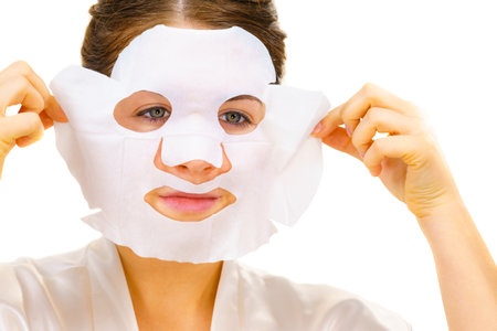 Woman applying sheet mask on her face, on white. Girl taking care of skin complexion. Beauty treatment. Skincare.の写真素材