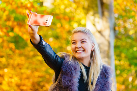 Blonde attractive woman wearing fashionable outfit artifice fur coat walking in autumn park, taking selfie photo with smartphone. Happy positive female having fun.の写真素材