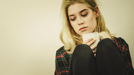 Sad contemplating woman sitting on couch alone drinking tea of coffee from mug.の写真素材