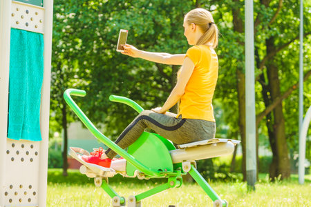 Young woman working out in the outdoor gym. Girl taking selfie photo with smartphone while doing exercises on street machine.の写真素材