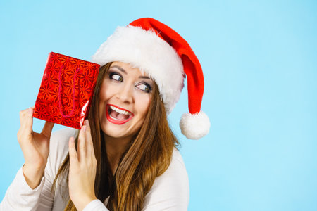 Smiling woman is happy to give Christmas gifts. Female wearing santa claus hat holding present red gift bag, on blueの写真素材