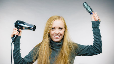 Female with blowing messy hair holding accessories hairdryer and brush. Blonde woman styling her very long disobedient hair.の写真素材