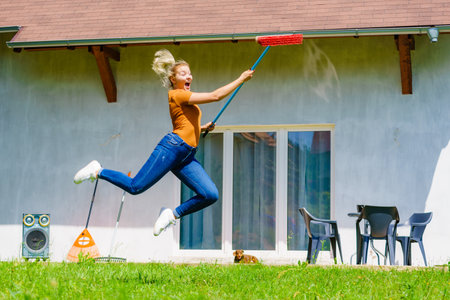 Happy woman working on her backyard, jumping with broom, red sweeping brush. Girl having fun, relaxing and enjoying life.の写真素材