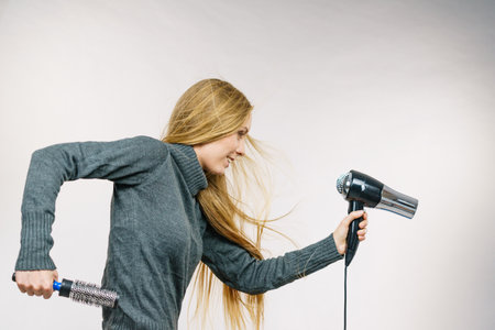 Female accessories holds hairdryer and round brush. Blonde woman styling her very long hair. side viewの写真素材
