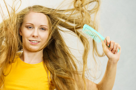 Blonde girl long tangled messy hair holds comb, untangling and brushing her hair. Haircare, treatment concept.の写真素材
