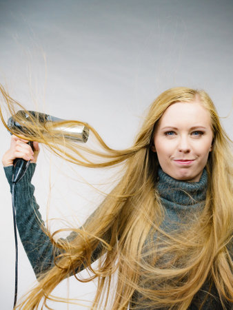 Blonde woman styling her very long hair. Female with blowing messy hair holding hairdryer. Hair accessories.の写真素材