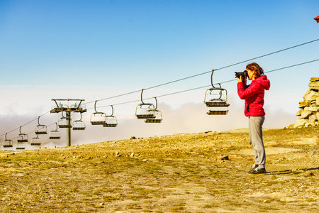 Tourist woman with camera taking travel photo in mountains. Empty chair lift in the distance. Torre mountain peak of Serra da Estrela, Portugal. Summer holiday trip.の写真素材