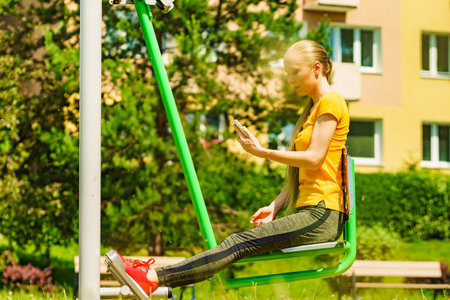 Young woman working out in outdoor gym. Girl holding mobile phone while doing exercises on street machine.の写真素材