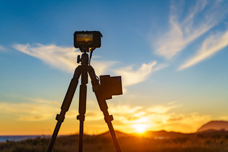 Professional camera on tripod taking picture film video of sunset over sea coast.