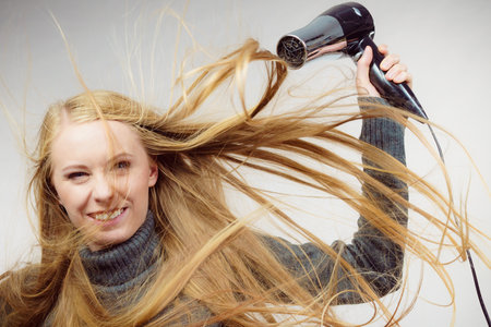 Blonde woman styling her very long hair. Female with blowing messy hair holding hairdryer. Hair accessories.の写真素材