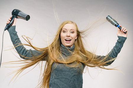 Female with blowing messy hair holding accessories hairdryer and brush. Blonde woman styling her very long disobedient hair.の写真素材