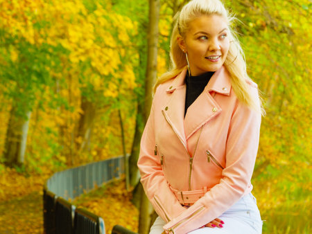 Blonde young woman wearing fashionable outfit. Happy female in pink leather jacket walking relaxing in autumn park.の写真素材