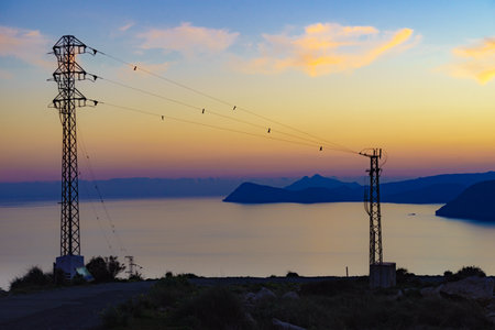 Coastline at sunset with electricity transmission pylons, power lines high voltage towers. Mesa...