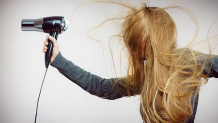 Blonde woman styling her very long hair. Female holding hairdryer. Hair accessories. Side viewの写真素材