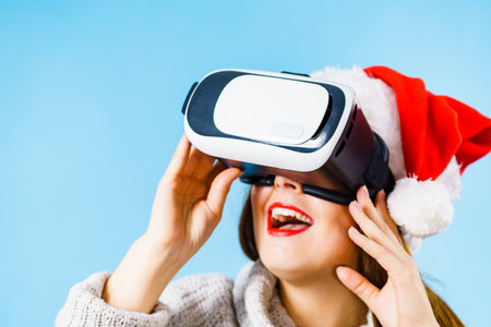 Woman wearing santa claus hat exploring space with virtual reality goggles headset. Emotional female watching 3d film tour in vr glasses box. New generation cyber christmas concept, on blue.の写真素材