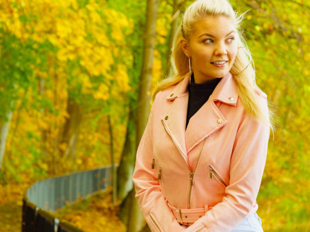 Blonde young woman wearing fashionable outfit. Happy female in pink leather jacket walking relaxing in autumn park.の写真素材