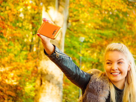 Blonde attractive woman wearing fashionable outfit artifice fur coat relaxing in autumn park, taking selfie photo with smartphone. Happy positive female having fun in sunny day.の写真素材