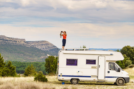 Woman enjoying trip with motor home, standing on roof of camper vehicle, holds camera, taking travel picture from mountain nature. Happiness, vacation and traveling.の写真素材