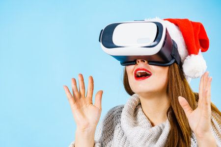 Woman wearing santa claus hat exploring space with virtual reality goggles headset. Emotional female watching 3d film tour in vr glasses box. New generation cyber christmas concept, on blue.の写真素材