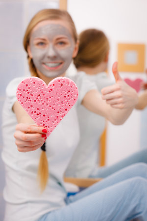 Facial dry skin and body care, complex treatment at home concept. Happy young woman loving having gray mud mask on her face, holding sponge in heart shape.の写真素材