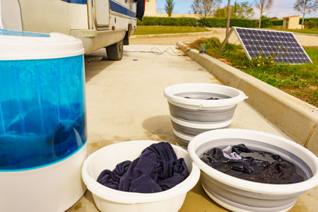 Doing laundry on vacation trip. Bowls with clothes and washing machine working. Caravan vehicle in the background. Camping on campsite.の写真素材