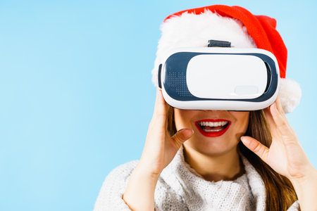 Woman wearing santa claus hat exploring space with virtual reality goggles headset. Emotional female watching 3d film tour in vr glasses box. New generation cyber christmas concept, on blue.の写真素材