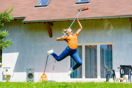 Happy woman working on her backyard, jumping with broom, red sweeping brush. Girl having fun, relaxing and enjoying life.の写真素材