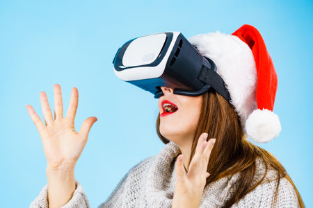 Woman wearing santa claus hat exploring space with virtual reality goggles headset. Emotional female watching 3d film tour in vr glasses box. New generation cyber christmas concept, on blue.の写真素材