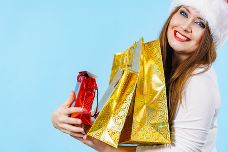 Young adult woman is happy to give Christmas gifts. Female wearing red Santa Claus hat holding paper bags presents. Blue backgroundの写真素材