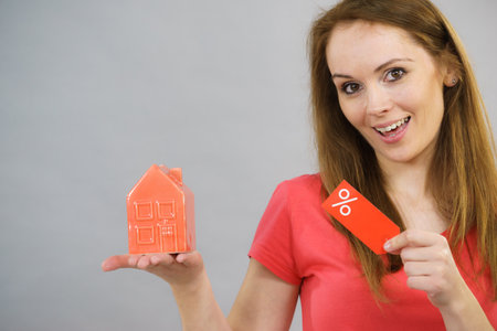 Woman is happy about buying house at affordable price. Female holding small red home model and sale percentage sign. Reduced housing prices. Fall and crisis of real estate market. Low rentの写真素材