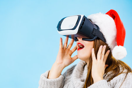 Woman wearing santa claus hat exploring space with virtual reality goggles headset. Emotional female watching 3d film tour in vr glasses box. New generation cyber christmas concept, on blue.の写真素材