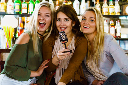 Three happy women having girls night out in pub, enjoying free time in karaoke bar sitting together holding old fashioned vintage microphoneの写真素材