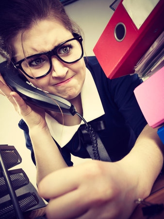 Angry businesswoman talking on phone sitting working at desk full of documents in binders.の写真素材