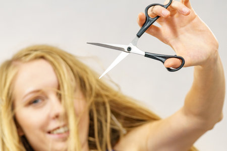 Young woman holding shears tool ready to hair trimming. Blonde female with scissors creating new hairdo coiffure. Hairstyle and haircut.の写真素材