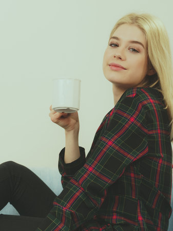 Pretty hipster young adult teenage woman sitting on sofa enjoying her leisure time drinking coffee...