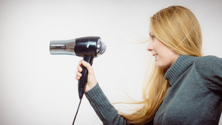 Blonde woman styling her very long hair. Female holding hairdryer. Hair accessories. Side viewの写真素材