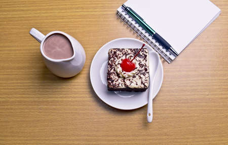 Chocolate cake with cherry and milk jug, notebook, cactus on wooden table