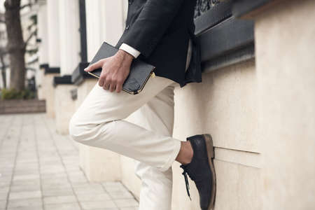 Elegant man wearing suit with black leather notebook in his hand is posing against a wall in the city street. Fashion image of a young businessman.の写真素材