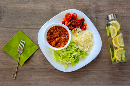 Top view on a bowl of hot ragout with meat, paprika and beans, stewed in tomato sauce and served with cherry tomatoes, cheese, fresh green cabbage on white plate on wooden background, next to a fork.の写真素材