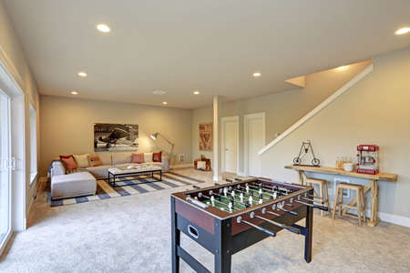 Light entertainment/game room design in a luxury new construction home with a wooden mini football table and soft beige sectional with red pillows offering comfortable seating atop a striped rug.の写真素材