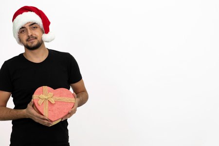 Young man wearing a Santa hat. He is wearing a black shirt. He is holding a gift box in the shape of a heart.の写真素材