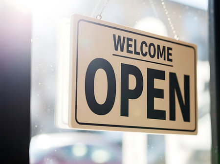 A beige rectangular sign with black text reading "WELCOME OPEN" hangs on a glass door. Sunlight streams in from the left, creating lens flare and illuminating dust particles.の素材
