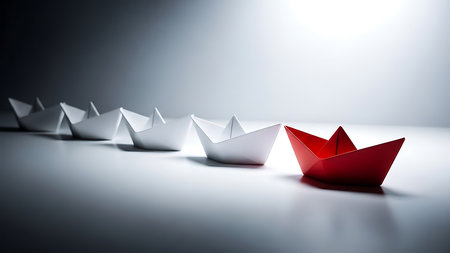 A single red origami paper boat leads a line of white origami paper boats across a smooth, reflective surface under dramatic lighting.の素材