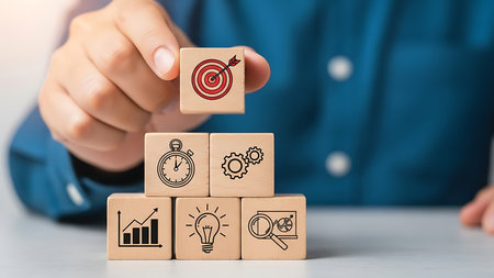 A hand places a wooden cube with a red and white target icon on top of a stack of wooden blocks featuring business-related icons like a stopwatch, gears, graph, lightbulb, and magnifying glass.の素材