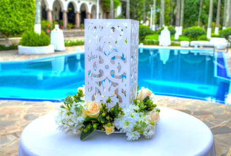 White large cubic candle holder decorated with flowers on its base, on a tall thin table at a celebration, on an overcast day. Outskirts of Quito, Pichincha, Ecuador.の写真素材