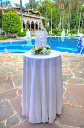 White large cubic candle holder decorated with flowers on its base, on a tall thin table at a celebration, on an overcast day. Outskirts of Quito, Pichincha, Ecuador.の写真素材