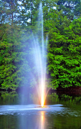 Water fountain spitting water out and up into the air. The fountain is part of a small lake surrounded by a forest in a park, Tampa, Florida, USAの写真素材