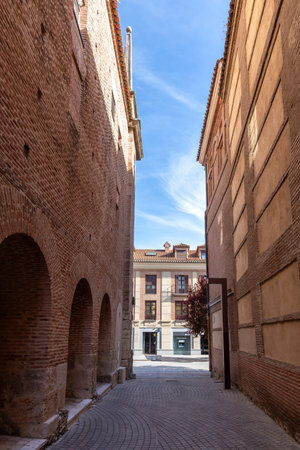 A narrow, traditional street in AlcalÃ¡ de Henares, walled by towering red brick facades, opens to reveal a building with balconies at the end under a clear blue sky.の写真素材